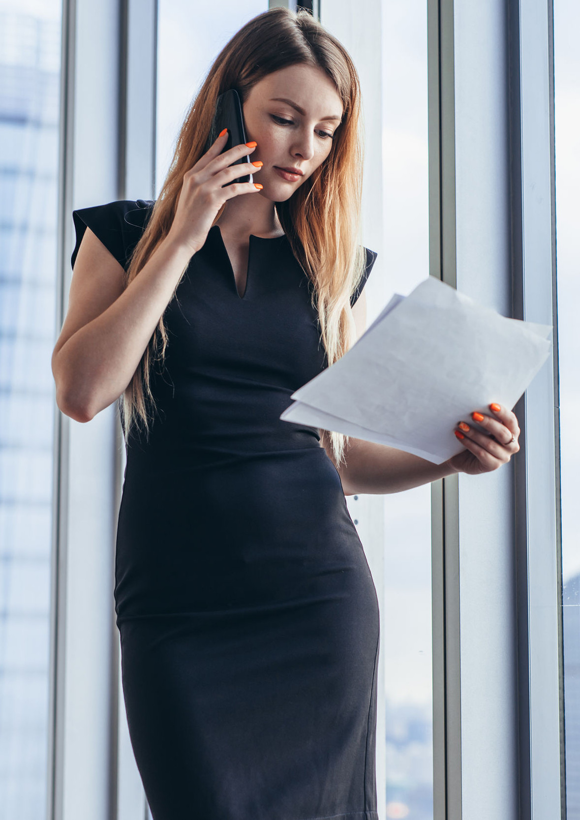 Elegant young female business owner holding documents talking on mobile phone discussing contract conditions standing in office.